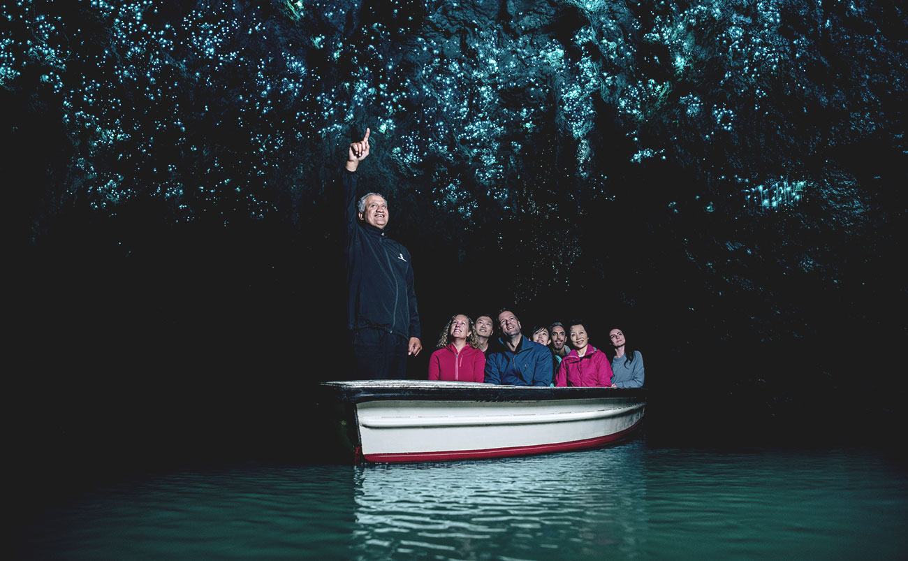 a man standing at the front of a small boat points to the roof of a cave illuminated by the glow of thousands of glow worms as guests watch on