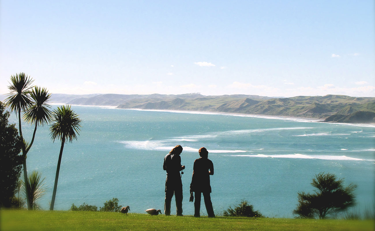 Two people are seen in silhouette as they stand on a green pasture in front of a view of the ocean and black sand beaches of Raglan, New Zealand