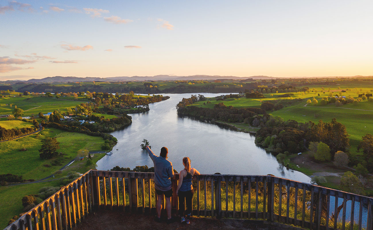 A man and a woman stand on a balcony overlooking the Waikato River as evening sun touches the riverbanks on either side of the river