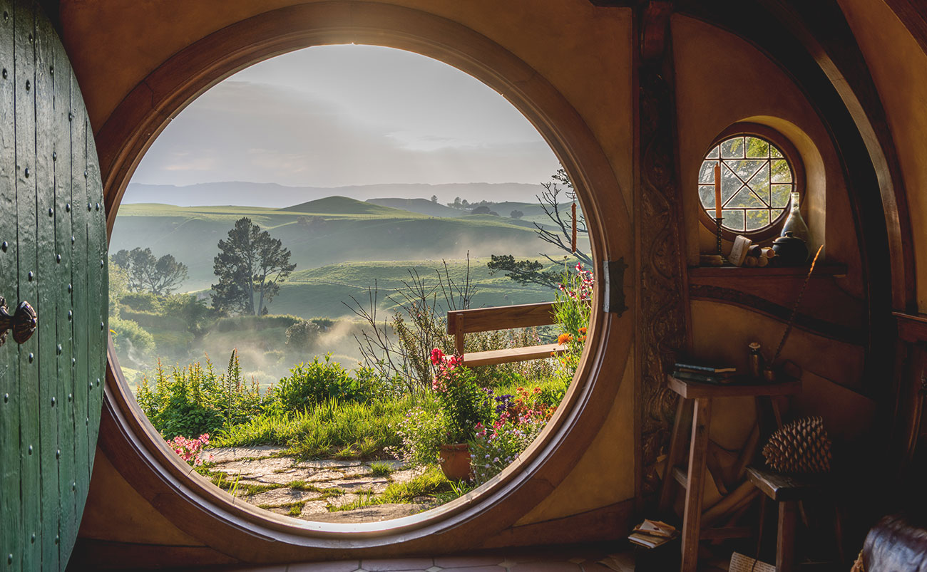 A view over the Hobbiton™️ Movie Set is seen through the circular door of a hobbit hole as morning mist envelops green rolling hills in the near distance